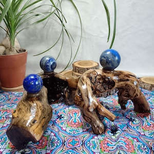 May include: Three wooden sculptures with polished, natural wood grain, each supporting a blue sphere. The sculptures are arranged on a colourful patterned fabric. A potted plant is visible in the background, adding a natural element to the composition.