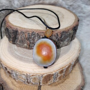 Puede incluir: Un collar con colgante que presenta una piedra lisa de forma ovalada con un núcleo central naranja rodeado por un anillo blanco y marrón. La piedra está suspendida de un cordón negro fino que incluye una pequeña cuenta texturizada color ámbar.