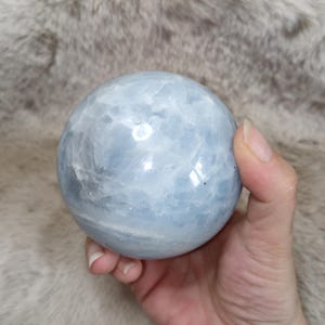 May include: A polished, spherical celestite crystal with a smooth, light blue surface featuring natural internal fractures and white mineral inclusions. The stone displays a glossy finish that reflects light, showcasing its translucent, pale blue color.