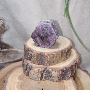 Puede incluir: Una gema de rubí natural en bruto, de color rosa intenso, colocada sobre tres rodajas de madera rústica apiladas. El rubí muestra variaciones de color y textura, con toques de inclusiones verdes más oscuras.