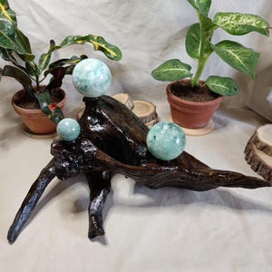 Pode incluir: Uma escultura de madeira flutuante polida marrom escuro com três esferas salpicadas de verde claro. A escultura é exibida em uma superfície neutra, com plantas em vasos ao fundo.