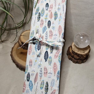 Op de afbeelding: Een rechthoekige, crèmekleurige stoffen etui met een herhalend patroon van kleurrijke veren in tinten blauw, roze en zwart. De etui is vastgebonden met een bijpassend stoffen lint. De etui is waarschijnlijk ontworpen om pennen of kunstbenodigdheden in te bewaren.