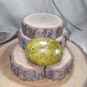Peut inclure: Une pierre verte polie de forme ovale avec une surface texturée, posée sur une pile de trois tranches de bois. La pierre a un aspect moucheté avec des nuances de jaune et de brun.