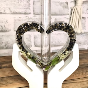 Könnte beinhalten: Eine klare Glas-Blumenknospenvase mit einem herzförmigen Harzrahmen, gefüllt mit getrockneten Blumen und Blättern. Die Vase wird von einem weißen, handförmigen Sockel getragen. Das Harzherz ist mit dunkelgrünen und hellgrünen getrockneten Blumen gefüllt.