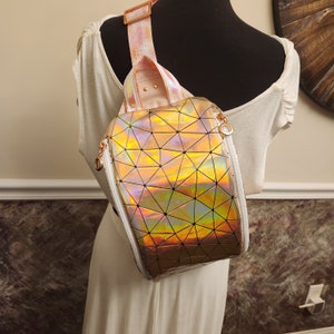 May include: A holographic iridescent pink and gold geometric pattern backpack with a white strap. The backpack has a round shape and a zipper closure.