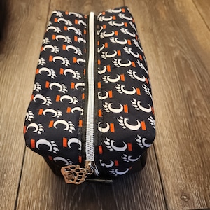 May include: Black zippered travel pouch with a white and orange paw print pattern. The zipper has a gold pull with a paw print design.