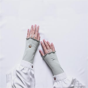 May include: Light green fingerless gloves with a ribbed texture and a small, decorative button. The gloves have a ruffled edge and a loop for the middle finger. The sleeves are long and extend up the arm.