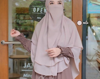 May include: A light brown hijab with a long, flowing design. The hijab is worn over a brown dress with ruffled sleeves. The text "Bergo Humaira" is visible at the bottom of the image.