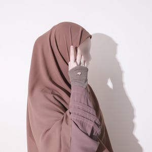 May include: A modest, brown hijab and matching sleeve cuffs. The cuffs have a ribbed texture and a decorative button. The person is making a peace sign with their fingers. The background is a plain white wall.