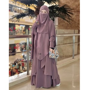 Puede incluir: Una abaya larga de color malva con un niqab a juego y una falda con volantes. La abaya tiene mangas largas con puños de botón y un detalle decorativo en la parte delantera. La mujer lleva un pequeño bolso de mano con estampado floral.