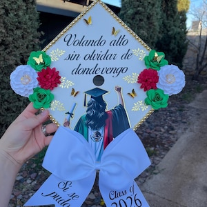 May include: A white graduation cap decorated with gold trim, paper flowers, and butterflies. The cap features the text "Volando alto sin olvidar de donde vengo" and a bow with "Si se pudo" and "Class of 2026".