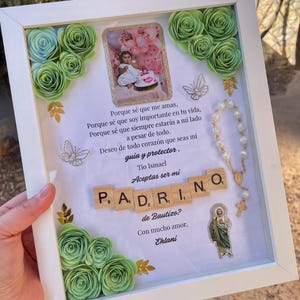 May include: A white shadow box frame containing a photograph of a child, light green paper roses, two silver butterfly charms, a white pearl rosary, a religious sticker, and wooden letter tiles spelling PADRINO, with Spanish text printed on the center.