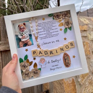 May include: A white shadow box frame containing a photograph of a baby, a rosary, paper flowers, gold stars, and wooden letter tiles spelling PADRINOS. The text reads: Mamá y Papá me criarán fuerte y siempre me mantendrán fiel, ¿Quieren ser mis Padrinos de Bautizo? Con Amor, Thiago Orozco.
