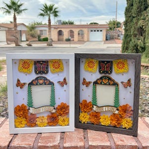 May include: Two shadow box frames, one white and one dark wood, each containing a central photo opening surrounded by paper flowers in orange and yellow, monarch butterflies, and a banner with the text AMOR ETERNO.