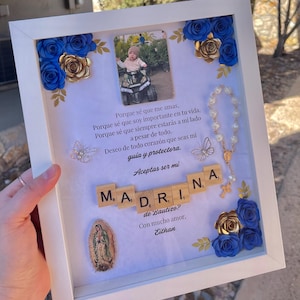 May include: A white shadow box frame containing a photograph of a child on a toy vehicle, blue and gold paper flowers, two silver butterfly charms, a white bead rosary, a religious medallion, and wooden letter tiles spelling MADRINA.
