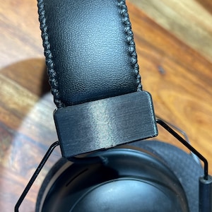 Peut inclure: Casque audio circum-auriculaire noir avec un bandeau et des oreillettes rembourrés. Le bandeau est cousu avec un fil noir. Le cadre métallique et les oreillettes sont noirs. Le casque est conçu pour l'écoute audio.