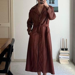 May include: A long-sleeved, ankle-length brown coat featuring a classic collar, a button-down front closure, and a matching fabric tie belt at the waist. The coat has a smooth texture, structured shoulders, and two side pockets, falling in a straight silhouette.
