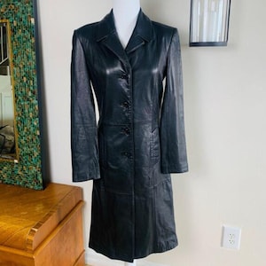 May include: A long black leather coat featuring a structured collar, long sleeves, and a front button closure. The coat has a fitted silhouette with visible seam detailing along the torso and waist, extending down to a knee-length hemline.
