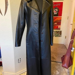 May include: A long black coat featuring a structured collar, long sleeves, and a front button closure with five visible buttons. The coat is displayed on a mannequin, showcasing a smooth finish and a tailored fit that extends to the lower calf area.