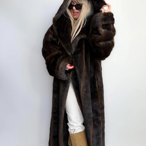 May include: A long, hooded faux fur coat in shades of brown. The coat has a floor-length design with a wide collar and sleeves. The fur appears soft and plush, creating a luxurious look. The coat is open in the front, revealing white trousers and brown boots.