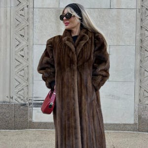 May include: A long, brown fur coat with a classic design. The coat has a wide collar and long sleeves. A red handbag is held in the hand. The background is a light-colored stone wall.