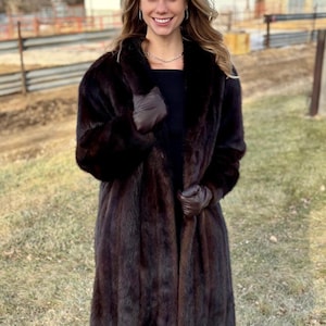May include: A long, dark brown fur coat with a wide collar. The coat is worn with brown leather gloves. The person is standing outside on a sunny day. The coat appears to be knee-length.
