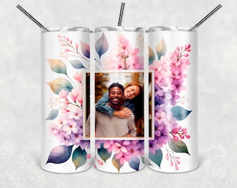 May include: A white cylindrical tumbler with a metal straw featuring a central rectangular photograph of a smiling couple. The design is surrounded by watercolor-style illustrations of pink and purple lilac flowers and soft green and blue leaves.