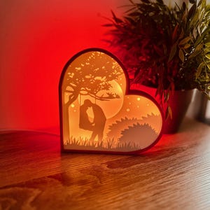 Peut inclure: Une lampe en papier en forme de cœur avec une silhouette d'un couple s'embrassant sous un arbre. La lumière chaude traverse les découpes complexes, créant une scène romantique. La lampe est posée sur une surface en bois.