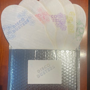 Puede incluir: Un conjunto de artículos blancos en forma de pétalo con diseños florales y el texto "DISCO DUSTERS" en varios colores, incluyendo azul, rosa, morado, amarillo y verde. Se muestran sobre un sobre de burbujas plateado con una etiqueta a juego.