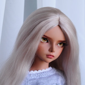 Puede incluir: Una muñeca con el pelo largo, liso y rubio platino que lleva un jersey gris con un gráfico de gato blanco y negro y el texto "I you think".