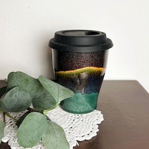 Könnte beinhalten: Ein Keramik-Reisebecher mit schwarzem Silikondeckel, der eine Farbverlaufglasur aufweist, die von einem dunklen, glitzernden Schwarz oben zu einem leuchtend gelben Band, gefolgt von tiefem Blau und einer soliden blaugrünen Basis mit glatter Oberfläche übergeht.