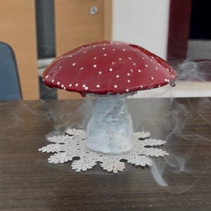 Könnte beinhalten: Eine skulptierte Pilzfigur mit einem glänzenden roten Hut, der kleine weiße Punkte aufweist, und einem dicken, strukturierten weißen Stiel. Der Pilz steht auf einer weißen, runden, filigranen Spitzendeckchen, umgeben von wirbelnden weißen Rauchschwaden.