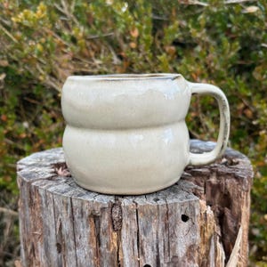 Könnte beinhalten: Handgefertigte, beige Keramiktasse mit einer einzigartigen, abgerundeten Form. Die Tasse hat einen Henkel und einen etwas dunkleren Rand. Die Tasse steht auf einer verwitterten Holzoberfläche, mit einem verschwommenen grünen Hintergrund.