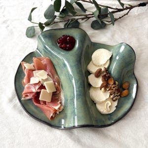 Op de afbeelding: Een turquoise keramische serveerschaal met drie secties. Eén sectie bevat prosciutto en kaas, een andere gesneden mozzarella en noten, en de derde bevat een klodder rode jam.