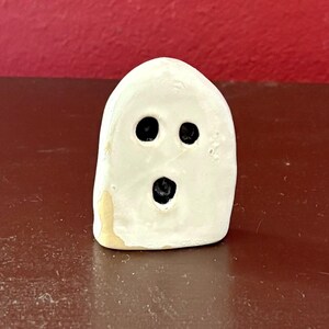 Könnte beinhalten: Eine kleine, weiße, abgerundete Keramik-Geisterfigur mit glänzender Oberfläche. Sie hat drei dunkle, kreisförmige Vertiefungen, die zwei Augen und einen offenen Mund darstellen, sowie einen kleinen Fleck aus unglasiertem beigem Material.