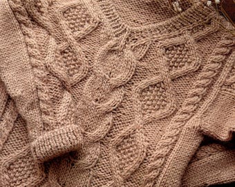May include: A light brown long-sleeved jumper featuring a prominent cable knit pattern with vertical braided textures and diamond-shaped sections. The garment has a ribbed crew neckline and ribbed cuffs on the sleeves, showcasing a thick, chunky knit construction.