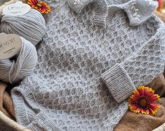 May include: A light grey knitted long-sleeve bodysuit with a textured diamond pattern, a collared neckline featuring white embroidered leaf details, and ribbed cuffs. Two balls of Cardiff Cashmere yarn are positioned next to the garment.