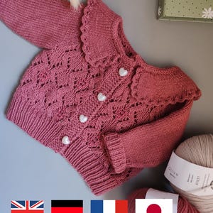 Puede incluir: Una rebeca de punto color rosa empolvado con cuello festoneado, mangas largas con puños acanalados y dobladillo acanalado. El cierre frontal consta de cuatro botones blancos en forma de corazón. El tejido presenta un diseño de rombos.
