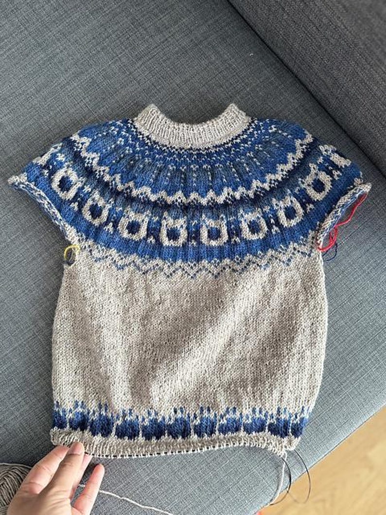 Puede incluir: Jersey de manga corta tejido a mano en tonos azules, grises y beige. El canes&uacute; presenta un detallado patr&oacute;n geom&eacute;trico en azul y gris. El cuerpo del jersey es de color beige liso, con un borde con estampado azul en la parte inferior.