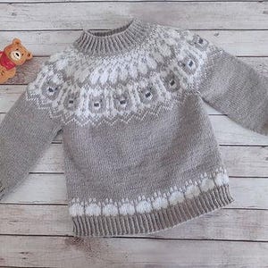 Puede incluir: Un su&eacute;ter de punto gris y blanco con canes&uacute; y pu&ntilde;os estampados. El su&eacute;ter presenta un cuello alto y acanalado y un dise&ntilde;o blanco y gris repetido en el pecho y el dobladillo inferior. Un peque&ntilde;o oso de peluche est&aacute; en la esquina superior izquierda.