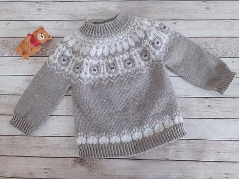 Puede incluir: Un su&eacute;ter de punto gris y blanco con canes&uacute; y pu&ntilde;os estampados. El su&eacute;ter presenta un cuello alto y acanalado y un dise&ntilde;o blanco y gris repetido en el pecho y el dobladillo inferior. Un peque&ntilde;o oso de peluche est&aacute; en la esquina superior izquierda.