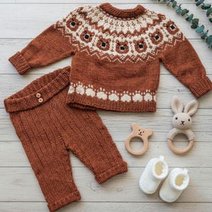 Puede incluir: Un conjunto de beb&eacute; de punto color &oacute;xido, que incluye un su&eacute;ter y pantalones, con un dise&ntilde;o de oso blanco y negro. El conjunto est&aacute; acompa&ntilde;ado de un mordedor de oso de madera, un conejito de peluche y botines blancos.