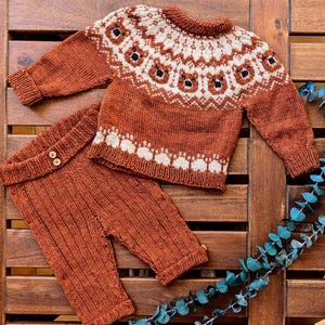 Puede incluir: Un conjunto de beb&eacute; de punto color &oacute;xido, que incluye un su&eacute;ter de manga larga y un pantal&oacute;n. El su&eacute;ter presenta un canes&uacute; color crema con un dise&ntilde;o de oso repetido y detalles en negro. El pantal&oacute;n tiene cierre de bot&oacute;n. El conjunto se muestra sobre una superficie de madera.