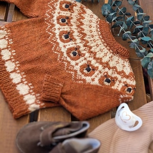 Puede incluir: Un su&eacute;ter de beb&eacute; tejido de color &oacute;xido con un patr&oacute;n geom&eacute;trico crema y negro. La imagen tambi&eacute;n muestra zapatos de beb&eacute; marrones, un chupete y un sombrero beige, todo dispuesto sobre una superficie de madera. El su&eacute;ter tiene mangas largas y cuello redondo.
