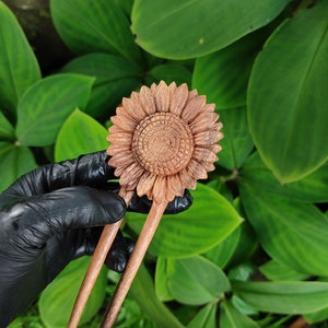 Può includere: Un fermacapelli in legno con un design a girasole intagliato. Il girasole è di colore marrone chiaro con dettagli di petali e centro intagliati. Il bastoncino ha due rebbi ed è posto su uno sfondo di foglie verdi.