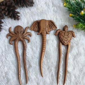 May include: Three wooden hair sticks carved with animal designs: an octopus, an elephant, and a turtle. The hair accessories are brown and have a smooth finish. The octopus has eight tentacles, the elephant has large ears, and the turtle has a shell.