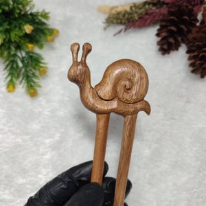 May include: A pair of wooden hair chopsticks with a carved snail design. The snail is brown and has a detailed shell. The chopsticks are a natural wood color.