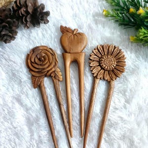 Op de afbeelding: Drie houten haarstokjes met gesneden bloemmotieven. De haarstokjes hebben een roos, een appel en een zonnebloem. Het hout heeft een natuurlijke bruine kleur. De haaraccessoires worden gepresenteerd op een witte, getextureerde ondergrond.