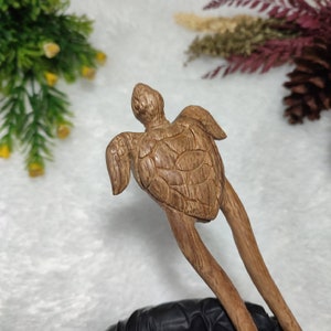 May include: A wooden hair stick featuring a carved turtle design. The turtle is light brown with detailed shell patterns and outstretched flippers. The stick has two long, slender prongs extending from the turtle's body.