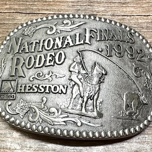 Op de afbeelding: Zilveren gesp met een rodeo-scène. Het toont een cowboy die een paard berijdt, een stier en de tekst "National Finals Rodeo 1992" en "Agco Hesston".